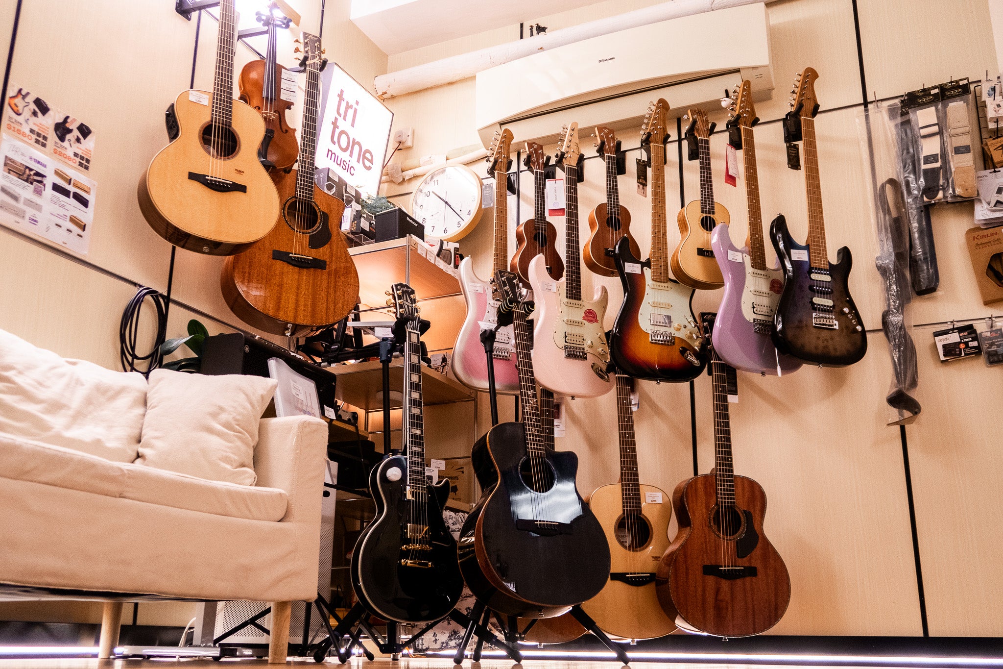 Guitars hanging on a wall in a music store with a couch and equipment in the foreground. Tritone Music 駿通音樂 租電子琴 租Band房 結他課程
